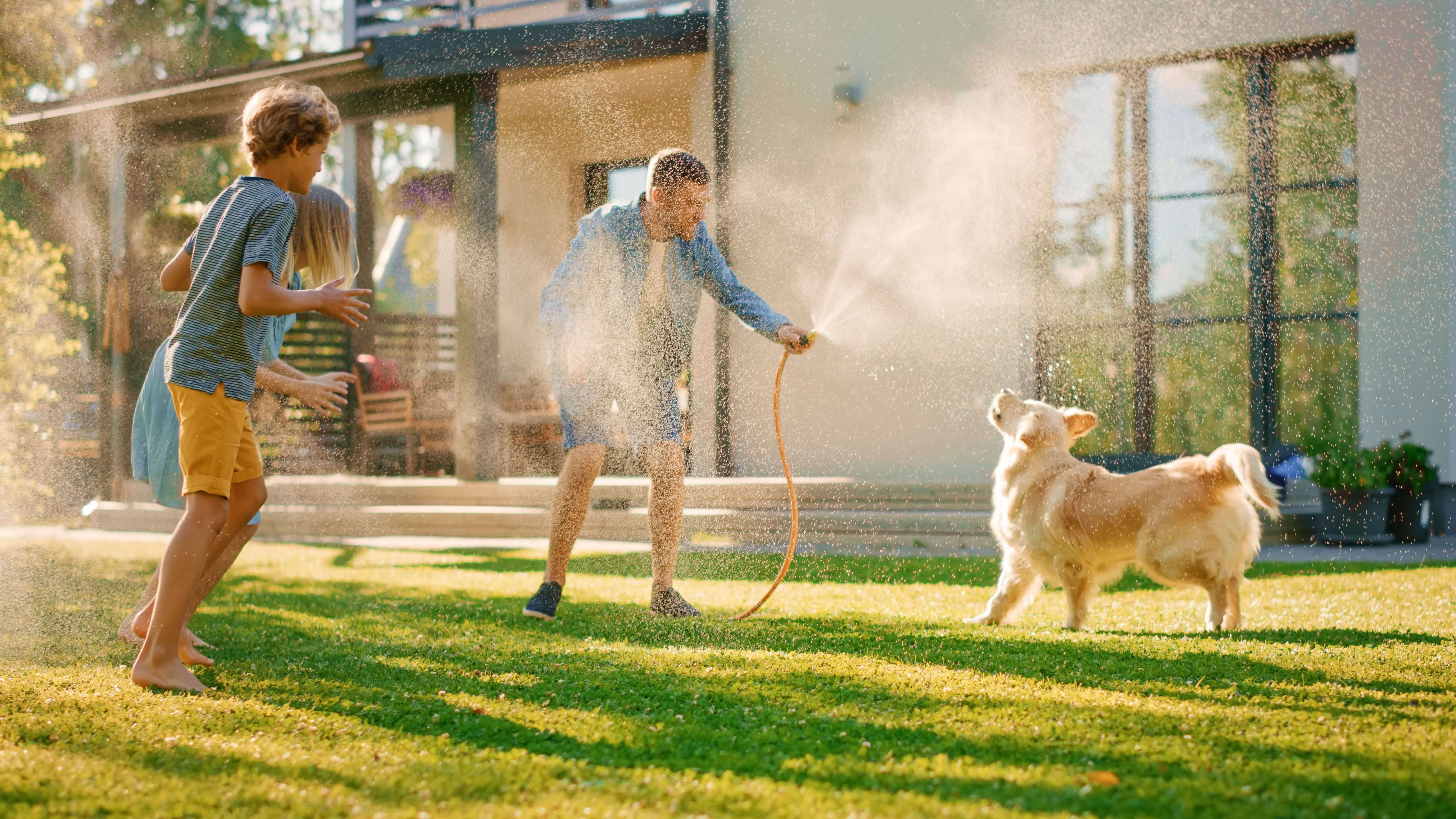 Een gezin speelt met de hond en de tuinslang op een groen grasveld voor een huis.